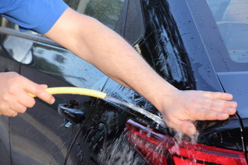 A white male hands washes a shiny black car rear fender and tail light with water from a hose close...