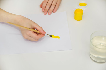 The child draws at a white table. Children's hands hold a brush and paint with yellow paint. Children's creativity.