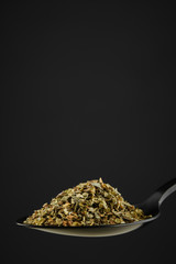 black spoon with Italian seasoning - dried oregano with thyme, basil and vegetables. black background. closeup