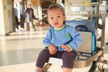 Little traveler, Cute Asian 2 years old toddler boy child with suitcase, sitting on trolley at airport, waiting for departure, Family travel & vacation with kid concept