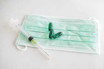 Green face mask with green capsule medicine and a hypodermic needle syringe on white table top.