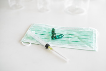 Green face mask with green capsule medicine and a hypodermic needle syringe on white table top.