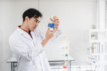 A male asian scientist staring seriously at a beaker with blue solution inside a laboratory setting.