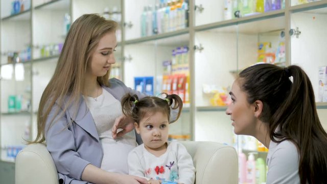 Young girl pharmacist giving advise to pregnant customer with cute baby girl. Pharmaceutical store concept
