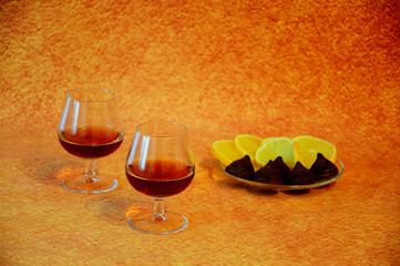 Two glasses with cognac and a plate with lemon slices and chocolate candies on a brown abstract...