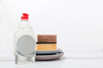Closeup of bottle of liquid detergent for cleaning, natural soap, sponge and stack of clean plates on the table against white wall.Empty space for text
