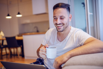 Fototapeta na wymiar Handsome smiling unshaven caucasian man in pajamas sitting in living room with laptop in lap and drinking his fresh morning coffee.