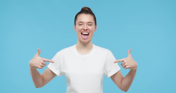Pretty girl pointing to her white t-shirt with fingers, showing empty space for your text or logo, isolated on blue background