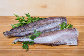 Uncooked defreezed hake carcasses among parsley on the wooden surface