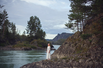  A girl in a white dress walks on a rocky shore near the river.