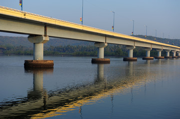 Fototapeta na wymiar Long road bridge over the river, side view. Powerful stone columns of bridge are reflected in the river water against the shore covered with green trees. Road bridge over Chapora river, Goa, India.