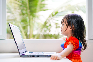 Adorable asian little girl is sitting and looking the laptop at home alone, concept of kid and technology in modern lifestyle and how to advice child to using the internet for learning and studying.