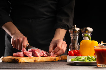 The chef cuts fresh pork for cooking. Against the background of vegetables. Kulinapiya, Restaurant business, recipes