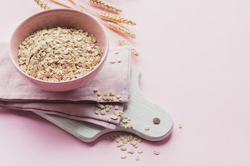 Bowl of dry oat flakes with ears of wheat on light pink background