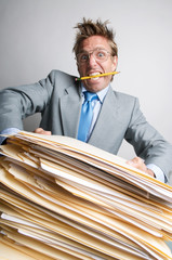 Stressed businessman with pencil in his mouth trying to lift a massive pile of paperwork on his desk