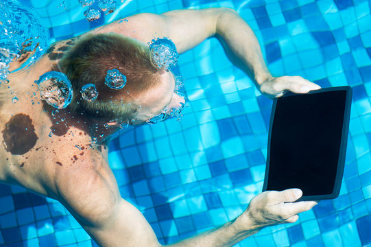 Man swimming underwater holding a digital tablet computer trailing bubbles in the pool behind him