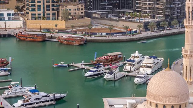 Yachts in Dubai Marina flanked by the Al Rahim Mosque and residential towers and skyscrapers aerial timelapse. Modern high rise skyline with boats and wooden dhows