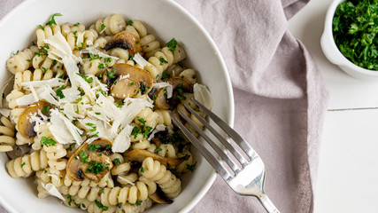 Pasta with mushrooms, parmesan and persil in white bowl on a grey napkin. There is some persil in a little bowl near. And you can see fork in a bowl. 