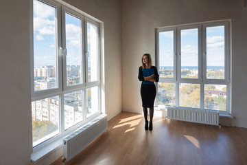 Girl realtor with a folder stands at the big windows of a spacious apartment