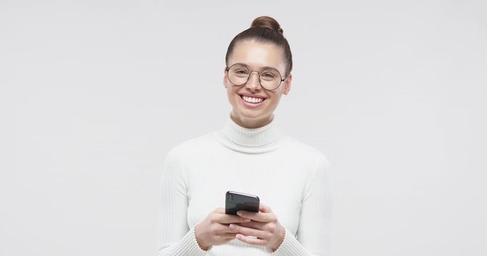 Young happy attractive european girl in white sweater smiling and laughing with smartphone in hands, isolated on studio gray background