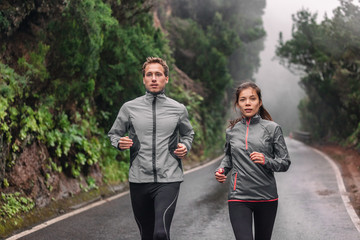 Running couple on wet park trail jogging in rain wearing sportswear jackets in cold weather. Asian woman, Caucasian man athletes training cardio.
