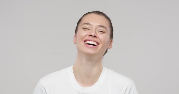 Happy cheerful young woman laughing out loud, looking at camera with joyful smile, isolated on studio gray background. Pretty student girl laugh. Slow motion