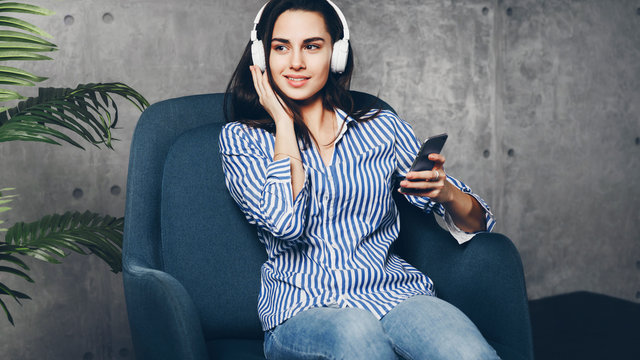 Young woman sitting in room and enjoying music streamed from mobile phone. Girl in earphones listening to songs from playlist in social media on her smartphone. Digital music solutions
