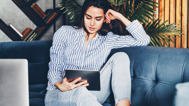 Young girl on sofa with digital tablet in her hand. Online communication via apps and messengers. Commenting and sharing in web media. Hipster girl checking her social network.