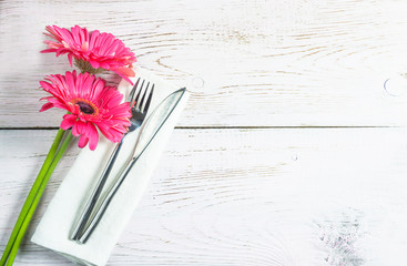 Festive table setting with handmade forged fork and knife on white napkin and bouquet of purple gerbera flowers on white paint wooden background. Space for text, flat lay