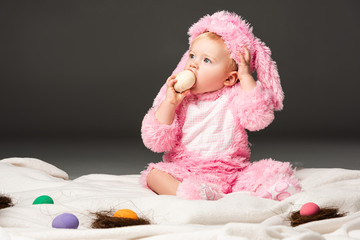 Child wearing rabbit costume, putting easter egg in mouth and sitting on blanket isolated on black
