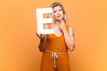 young latin pretty woman confused, doubtful, thinking, holding the letter E of the alphabet to form...