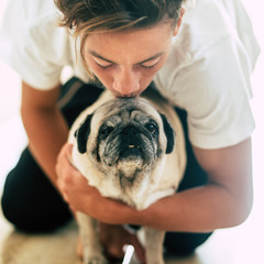 close up and portrait of two best friends at home together - teenager kissing the head of a pug - pet and domestic dog concept