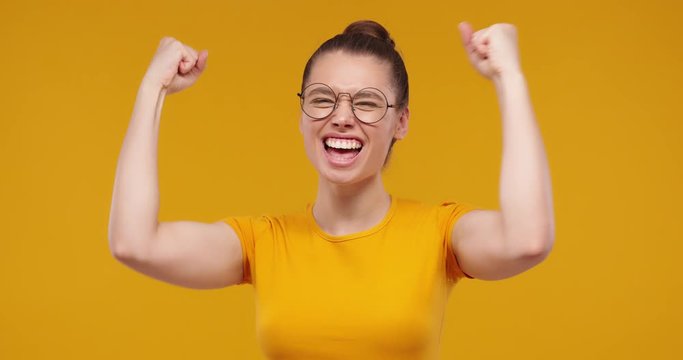 Young pretty fan girl shouting while her team win, raised both fists in victory gesture, isolated on studio yellow background