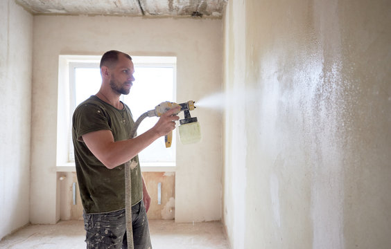 Concentrated worker painting with airbrush, apartment repairment against small window in the daytime. Man with a beard is dressed in paint-smeared T-shirt. Repair concept