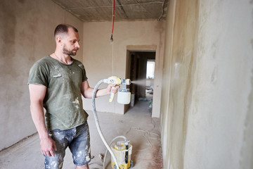 Male builder is painting with airbrush wall, house repairment. Large volume of work is visible in the background. Man with a beard is dressed in paint-smeared clothing. Repair concept