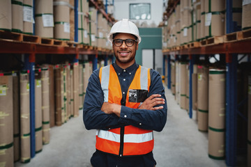 Portrait smiling worker wearing orange vest and white helmet with arms crossed in a large warehouse...
