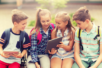 primary education, friendship, childhood, technology and people concept - group of happy elementary school students with backpacks sitting on bench and talking outdoors