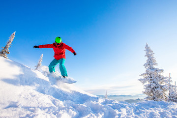 Young man snowboarder jump down slope mountains forest. Winter sport sun day, blue sky, fast speed dust snow
