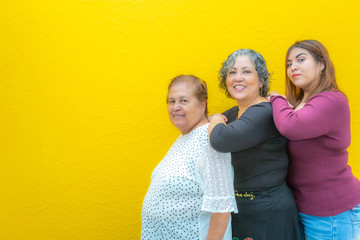 Grandmother, daughter and granddaughter in a row looking at the camera, three generations of Mexican women smiling in casual clothes on a yellow background, space for text