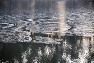 A thin crust of ice melts on the lake. Frosty pattern on the water in circles. The rays of the setting sun are reflected from the water.