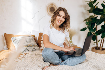 An amazing smiling girl in casual wear sits on a bed with a laptop and spends time on the Internet. Home leisure