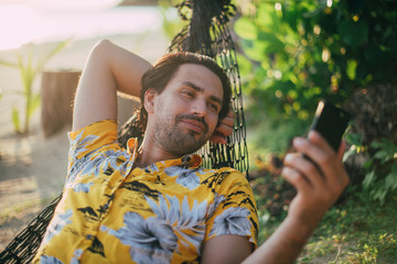 A young man lies in a hammock on a sunny beach with a phone.