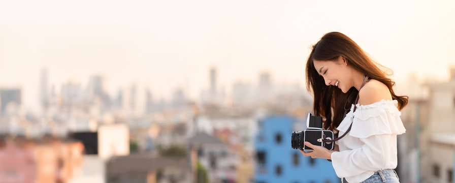 Portrait of Pretty Asian woman standing at outside while holding retro camera with Buildings background. Happy, Having Fun. Banner, Panoramic.