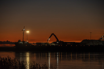 Fototapeta premium Silhouette of working crane in sea port near Parnu bay in cold winter evening against background of clear sunset sky. Port lights reflecting in water and reeds in foreground 