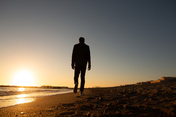 Fototapeta premium Young man and sunset on Mediteranean sea. One man walking on the beach and looking at the sunset over the sea. Solitary man. A colorful sunset sea, man observing the beauty of nature. 