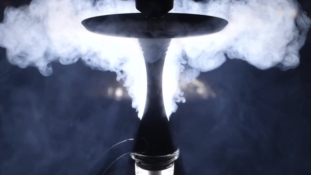 There is a hookah in a dark room. The hookah is illuminated at the back. They smoke hookah. Steam falls on the table and blows the hookah.