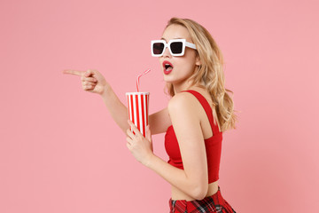 Side view of young woman in red sexy clothes, dark sunglasses isolated on pastel pink background. People lifestyle concept. Mock up copy space. Hold cup of cola or soda, pointing index finger aside.