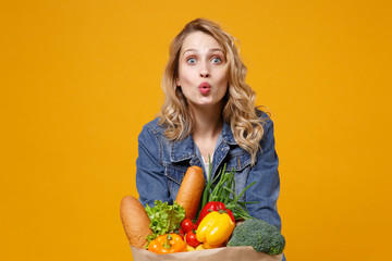 Pretty young woman in denim clothes posing isolated on yellow orange background. Delivery service from shop or restaurant concept. Hold brown craft paper bag for takeaway mock up with food products.