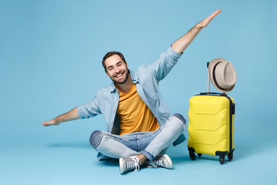 Smiling Traveler Tourist Man In Yellow Clothes Isolated On Blue Wall Background. Male Passenger Traveling Abroad On Weekend. Air Flight Journey Concept. Sit Near Suitcase, Spreading Hands Like Flying.