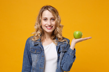 Smiling young woman in denim clothes isolated on yellow orange background studio portrait. Proper nutrition vegetarian food, healthy lifestyle diet meal. Diabetes dieting concept. Hold green apple.
